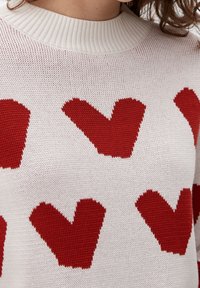 Stickad tröja i vitt med en ribbad krage som har växelvis röda hjärt-mönster över hela, som visar en texturerad och ventilerande tyg.