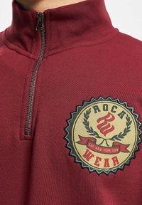 Burgundi pusiau užtrauktuko medvilninis džemperis su apskrito logotipo aplikacija, dekoratyviais elementais ir kontrastingomis siūlėmis aplink užtrauktuką.