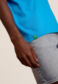 Felblauw katoenen T-shirt zichtbaar aan de onderkant met een groene label. Hand gedeeltelijk zichtbaar, die lichte denim shorts vasthoudt met een gladde textuur.