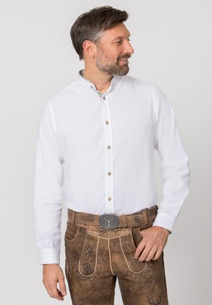 Mann trägt traditionelle braune Lederhosen aus Leder mit bestickten Mustern und einem weißen langärmeligen Hemd, schaut zur Seite.