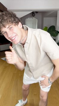 Beżowa koszula w paski z krótkim rękawem, białe szorty, srebrny zegarek oraz trampki z kokardą. Wnętrze z drewnianą podłogą i roślinami.