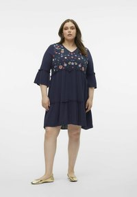 Vestido navy con bordados florales en el corpiño, escote en V, mangas acampanadas y volantes en capas; combinado con bailarinas doradas.