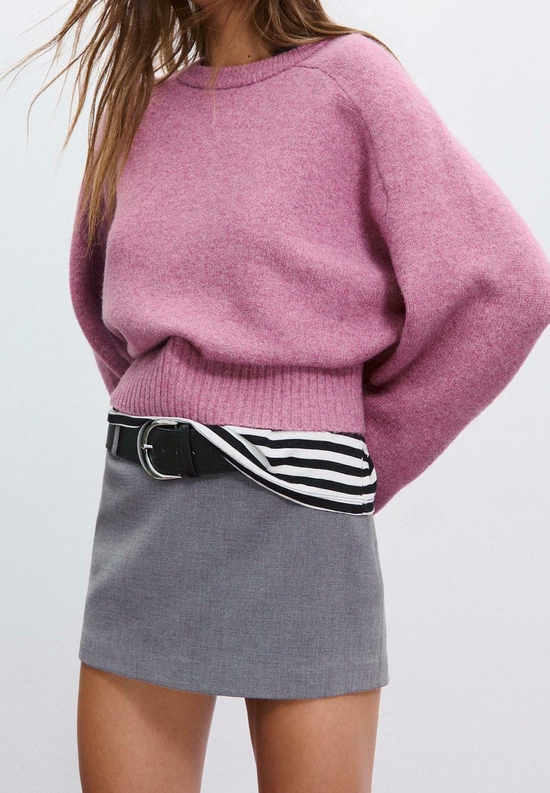 Femme portant un pull rose oversize, une chemise rayée noire et blanche rentrée, une mini-jupe grise, et une ceinture noire avec une boucle argentée.