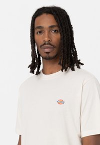 Camiseta de algodón color crema con un pequeño logo colorido en la parte izquierda del pecho, mangas cortas y diseño de cuello redondo.