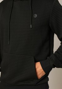 Haut à capuche noir avec un motif texturé et matelassé. Il dispose d'une poche kangourou à l'avant, d'une capuche avec cordon de serrage ajustable et de poignets côtelés.