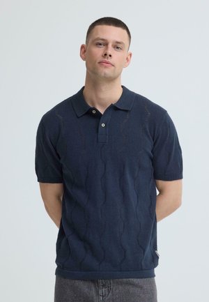 Jonge man met kort haar, gekleed in een donkerblauw, gestructureerd polohemd met korte mouwen, staand tegen een effen lichte achtergrond.