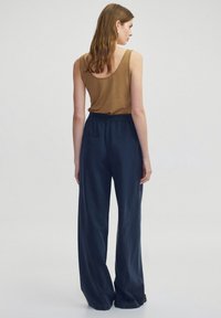 Bruine mouwloze top gecombineerd met donkerblauwe, wijde broek met een soepele textuur en een elastische tailleband, gezien vanuit de achterkant.