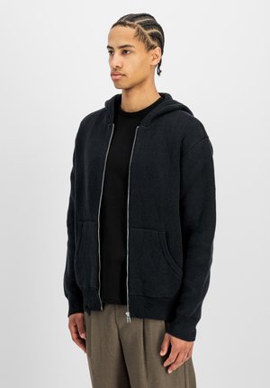 Giovane uomo con capelli intrecciati che indossa una felpa nera con zip sopra una camicia nera e pantaloni marroni, in piedi davanti a uno sfondo chiaro e uniforme.