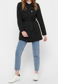 Schwarzer, doppelt geknöpfter Trenchcoat mit tailliertem Gürtel, ausgestattet mit einem Kragen und Knopfakzenten, kombiniert mit hellblauen Jeans und weißen Sneakers.