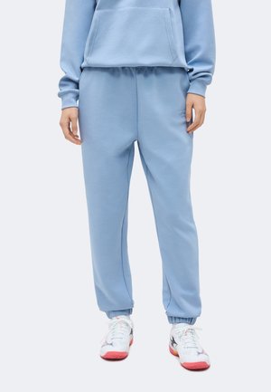 Osoba nosiaca svetlomodré tepláky a zodpovedajúcu mikinu s bielymi teniskami s červenými podrážkami a čiernymi detailmi.