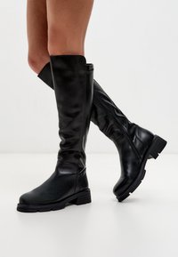 Botas de cuero negro hasta la rodilla con una textura lisa, punta redondeada, tacón bajo tipo bloque y detalle de cremallera lateral.