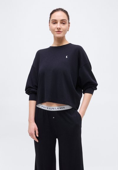 Νεαρή γυναίκα που φοράει ένα φαρδύ μαύρο πουκάμισο μακρυμάνικο και μαύρο παντελόνι με εμφανές λάστιχο μέσης "Polo Ralph Lauren", στέκεται μπροστά σε απλό φόντο.