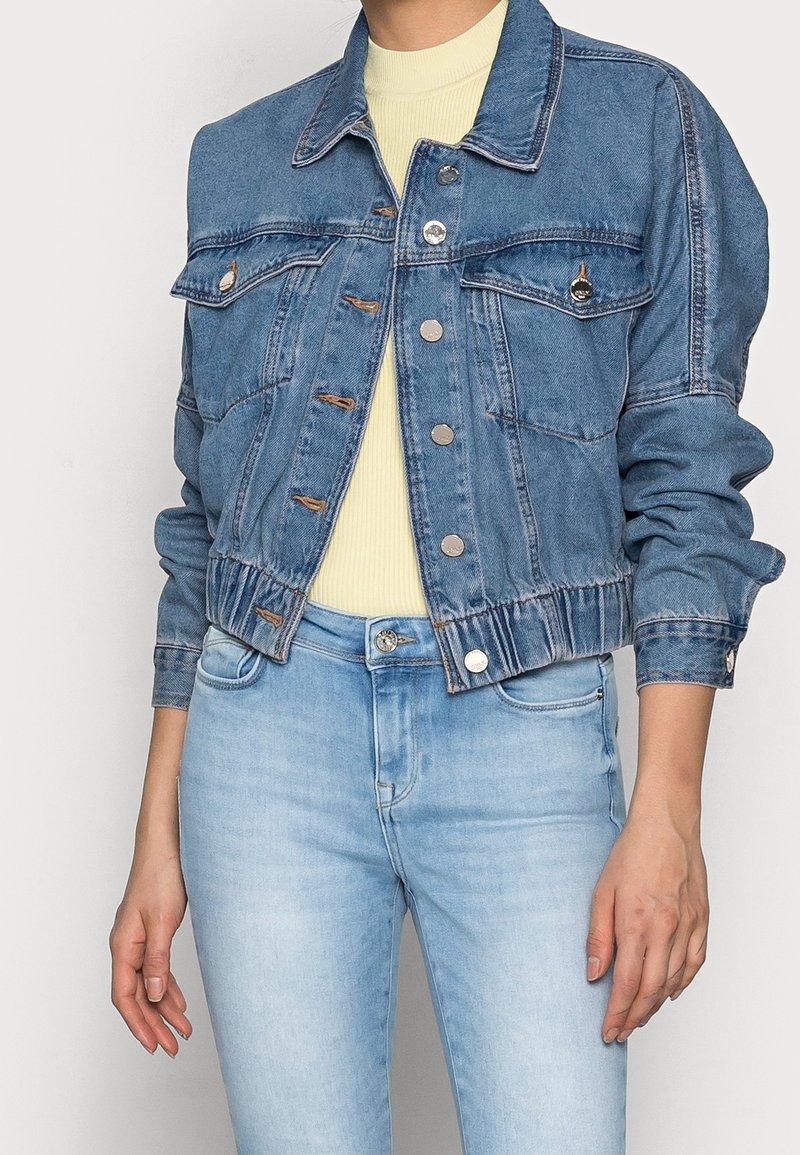 Femme portant une veste en jean bleue courte par-dessus un haut côtelé jaune pâle avec un jean en denim bleu clair, debout, les mains détendues sur les côtés.