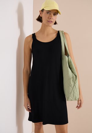 Femme portant une robe noire sans manches et une casquette jaune, portant un grand sac d'épaule matelassé vert clair.