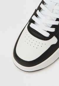 Pantof sport alb-negru cu partea superioară din piele, șireturi plate, vârf perforat și talpă de cauciuc. Cusături albe de-a lungul secțiunilor negre.