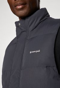 Dunkelgraue gepolsterte Weste mit hohem Kragen, die ein Logo auf der Brust und Druckknopfverschlüsse aufweist. Glatte Textur, minimalistisches Design.