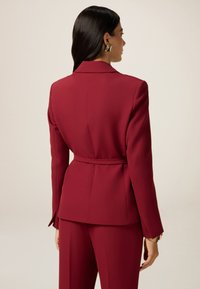 Blazer ajusté bordeaux avec une forme cintrée, taille ceinturée, épaules structurées et tissu lisse ; couture visible et revers élégant.