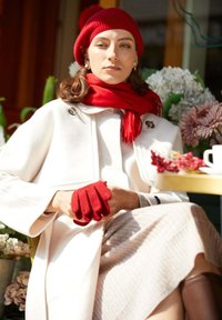 Cappotto di lana bianco con una sciarpa rossa a contrasto e guanti abbinati. Modella seduta con uno sfondo floreale, che mostra una postura rilassata.