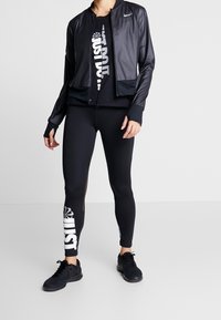 Czarna sportowa kurtka z kołnierzem na stójce i zapinana na zamek z przodu, zestawiona z czarnymi legginsami z białym logo i siateczkowymi akcentami.