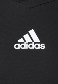 Logo Adidas s třemi bílými diagonálními pruhy nad názvem značky na černém texturovaném látkovém pozadí.