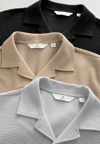Three collared shirts in black, beige, and light gray, featuring a textured ribbed pattern. Labels indicate brand and size information.