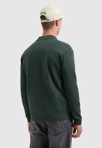Groene trui met kraag, gemaakt van textuurstof. Draag met grijze jeans en een lichte pet met borduursel aan de achterkant.