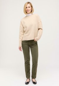 Béžový pletený svetr s žebrovanými manžetami, spárovaný s úzkými olivově zelenými džíny a černými plochými botami. Hladká textura a volný střih po celé délce.
