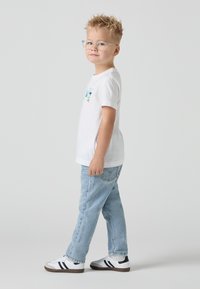 T-shirt blanc à manches courtes avec un motif coloré, jeans en denim bleu clair et baskets blanches avec des accents noirs. L'enfant porte des lunettes transparentes.