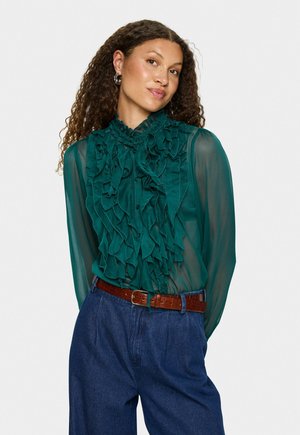 Blusa teal con volants, colletto alto e chiusura a bottoni, abbinata a pantaloni in denim blu a vita alta e una cintura marrone strutturata.