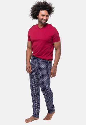 Mann mit lockigem Haar lächelt, trägt ein rotes kurzärmeliges Hemd und gemusterte marineblaue Pyjamahosen, steht barfuß vor weißem Hintergrund.