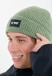 Grön stickad mössa med ribbad textur, med en svart rektangulär lapp märkt "SLOPE." Formad för att passa tätt på huvudet.