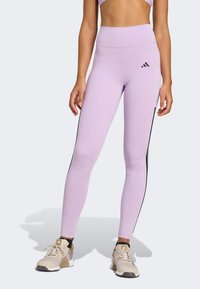 Leggings lilas clairs avec une taille haute, des bandes noires sur les côtés et un logo Adidas sur la cuisse gauche. Tissu lisse et extensible.