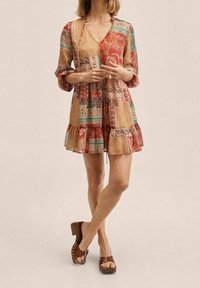 Gaun bermotif dalam nuansa hangat dengan hem berkerut, lengan tiga perempat, dan pinggang yang dipadatkan. Dipadukan dengan sepatu berhak tebal berwarna cokelat.