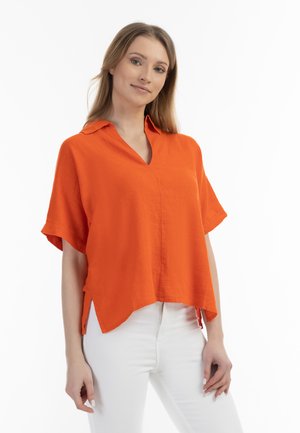 Kurzarmpullover aus orangefarbenem Leinen mit V-Ausschnitt, seitlichen Schlitzen und lockerer Passform, kombiniert mit weißen Jeans für den Kontrast.