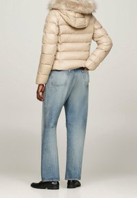 Beżowa kurtka puchowa z kapturem wyłożonym futerkiem, w zestawieniu z luźnymi niebieskimi dżinsami. Kurtka ma pikowany wzór i elastyczne mankiety.