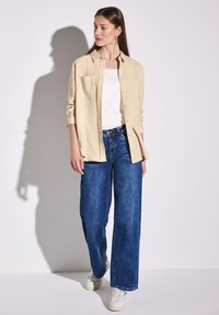 Béžová strukturovaná košile na knoflíky přes bílý top, v kombinaci s širokými modrými džíny a bílými teniskami. Košile má dvě kapsy na prsou.