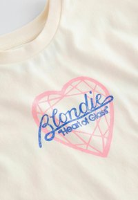 Kerman vaaleanpunaisella sydänkuviolla varustettu puuvillahihaton t-paita, jonka päällä on siniset tekstit "Blondie 'Heart of Glass'."