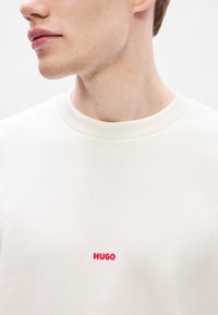 Λευκή φούτερ μπλούζα με στρογγυλή λαιμόκοψη και μικρό κόκκινο λογότυπο "HUGO" στο στήθος, κατασκευασμένη από ομαλή, υφή υφάσματος.
