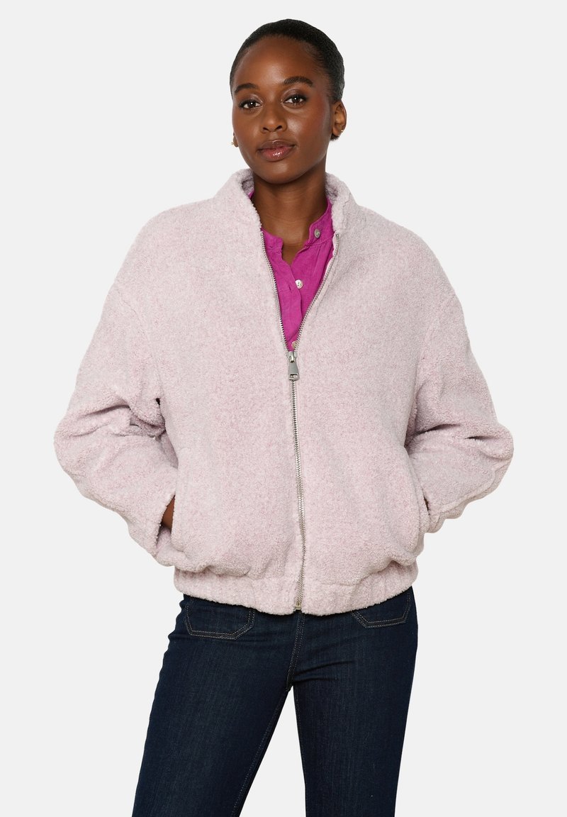 Šviesiai rožinė dirbtinio kailio striukė su užtrauktuku priekyje, raukšlėtomis rankovėmis ir elastinga juosmens juosta, derinama su magentos spalvos marškiniais ir tamsiomis džinsais.