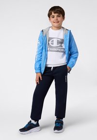 Gutt som har på seg en blå hettejakke, hvit Champion-genser, svarte joggebukser og marineblå sneakers, stående med en hånd i lommen.