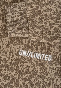 Krátká mikina v béžové barvě s hnědým puntíkovaným vzorem. Má žebrované manžety a na přední straně bíle potištěn nápis "UN//LIMITED".