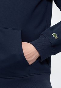 Main dans la poche avant d'un sweat-shirt bleu marine avec poignets côtelés et petit logo crocodile sur la manche.