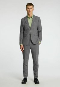 Grijze op maat gemaakte pakken met schouderkleppen, enkele knoopsluiting en vlakke broek. Draag het over een lichtgroen gestreept shirt. Zwarte schoenen.