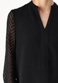 Musta pusero, jossa on halkio V-pääntie ja läpinäkyvät pitkät hihat, joissa on teksturoitu sahalaitakuosi ja pieniä kohotettuja pisteitä.