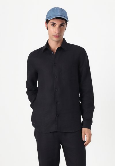 Chemise noire à manches longues en tissu léger et texturé, avec un col à boutons et une poche poitrine. Portée avec une casquette en denim bleu.