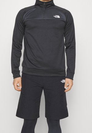 Muž nosící černý pulovr s dlouhým rukávem a polovičním zipem a kraťasy po kolena s logem The North Face na hrudi a stehně.