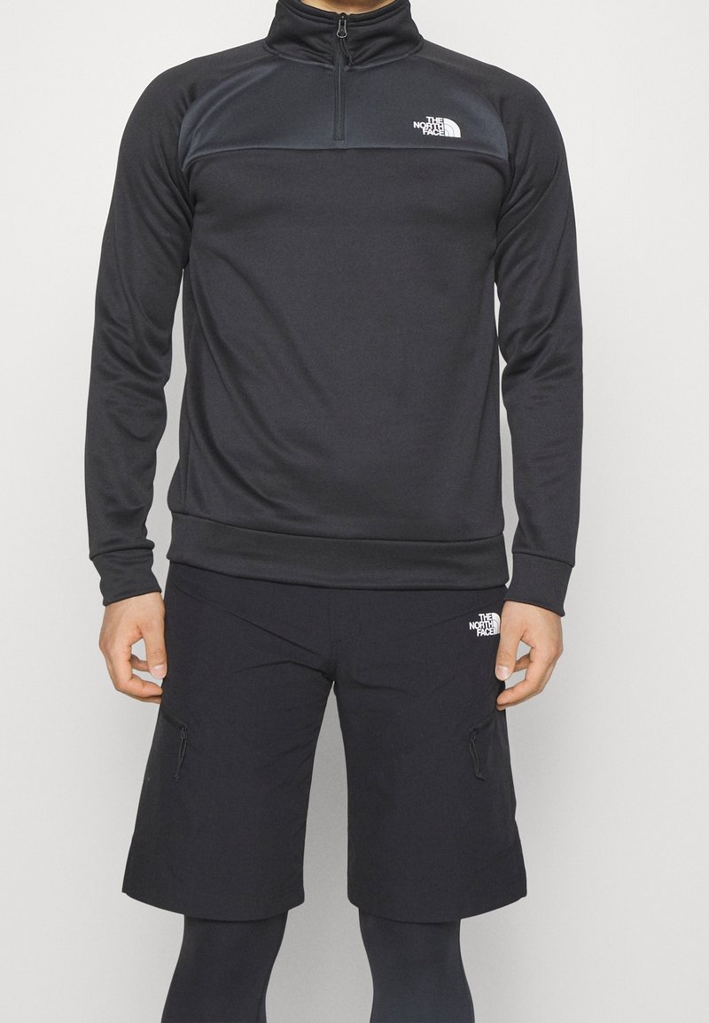 Homme portant un pull noir à manches longues avec une fermeture éclair jusqu'à mi-hauteur et un short jusqu'aux genoux avec le logo The North Face sur la poitrine et la cuisse.