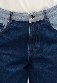 Τζιν παντελόνι με σκούρο πλύσιμο, με ανοιχτό μπλε ζώνη μέσης, ραφές σε χάλκινο χρώμα και μπροστινές τσέπες. Κουμπί με το λογότυπο στη μέση.