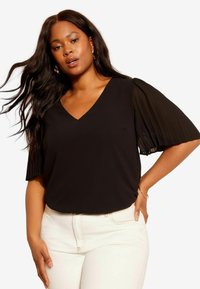 Un haut noir avec un col en V et des manches courtes plissées transparentes, associé à un pantalon blanc ajusté. Texture lisse et design minimaliste.