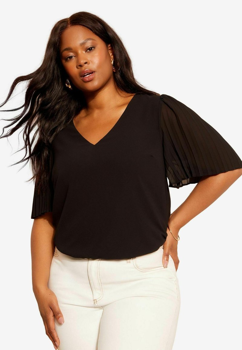 Un haut noir avec un col en V et des manches courtes plissées transparentes, associé à un pantalon blanc ajusté. Texture lisse et design minimaliste.